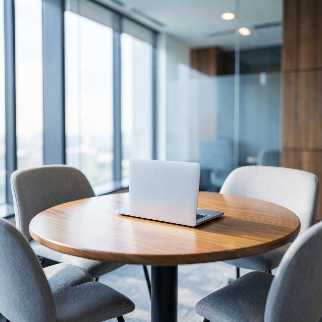 An unbranded open laptop on a round wooden table surrounded by empty upholstered chairs in a bright office.