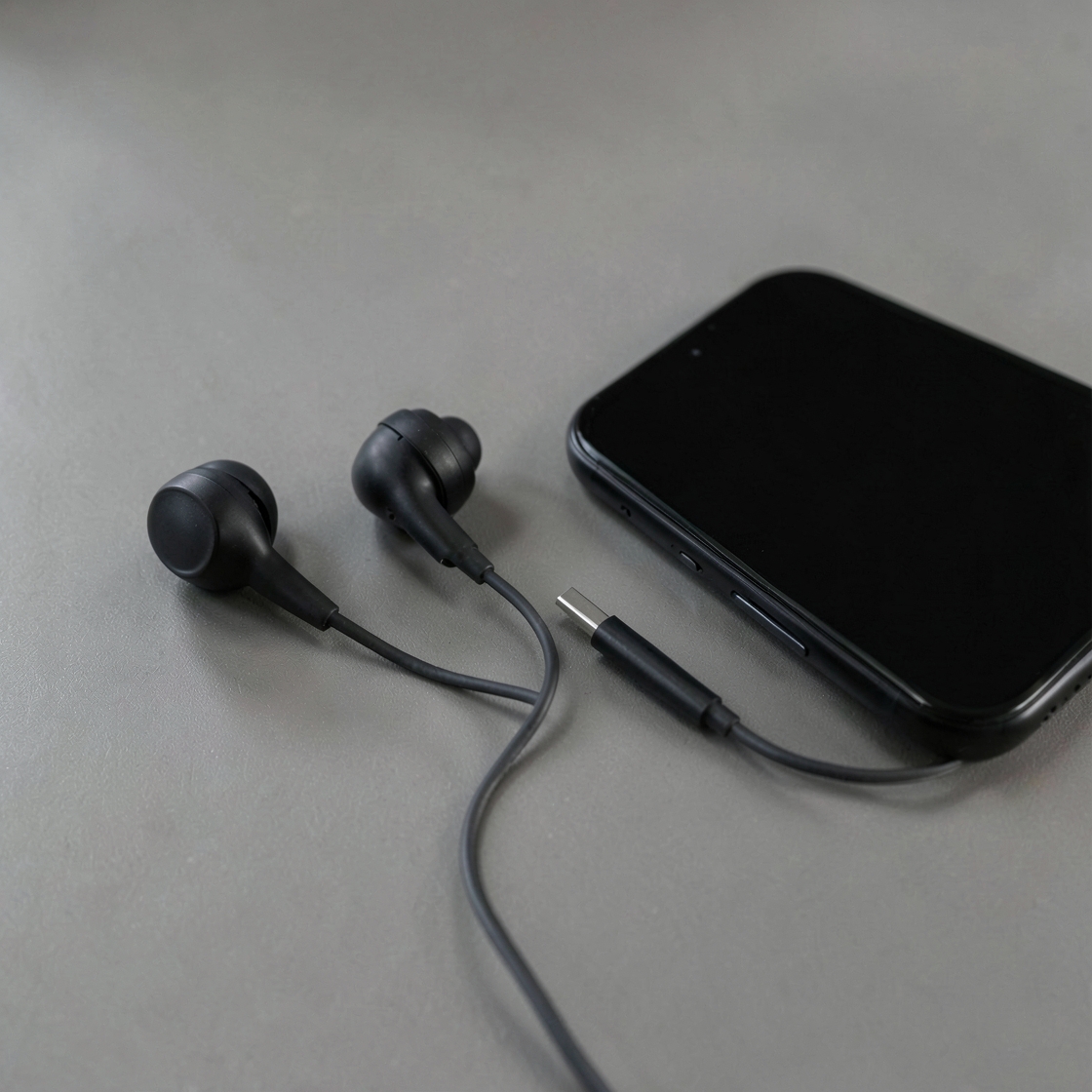 Two unbranded black wired earbuds and a USB-C plug beside a dark-screen phone