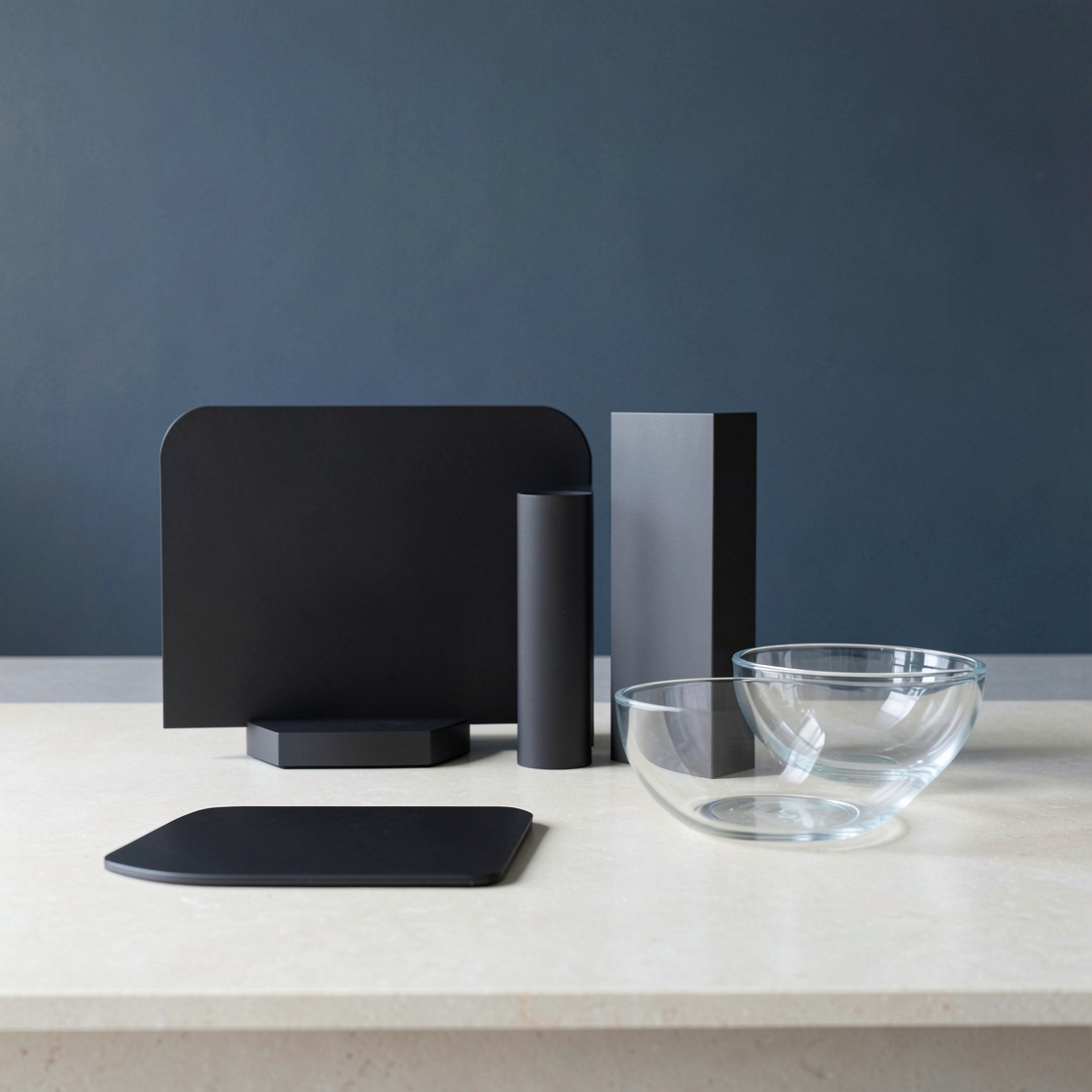 Blank dark panel, upright geometric columns, a flat slab and clear glass bowls on a pale table