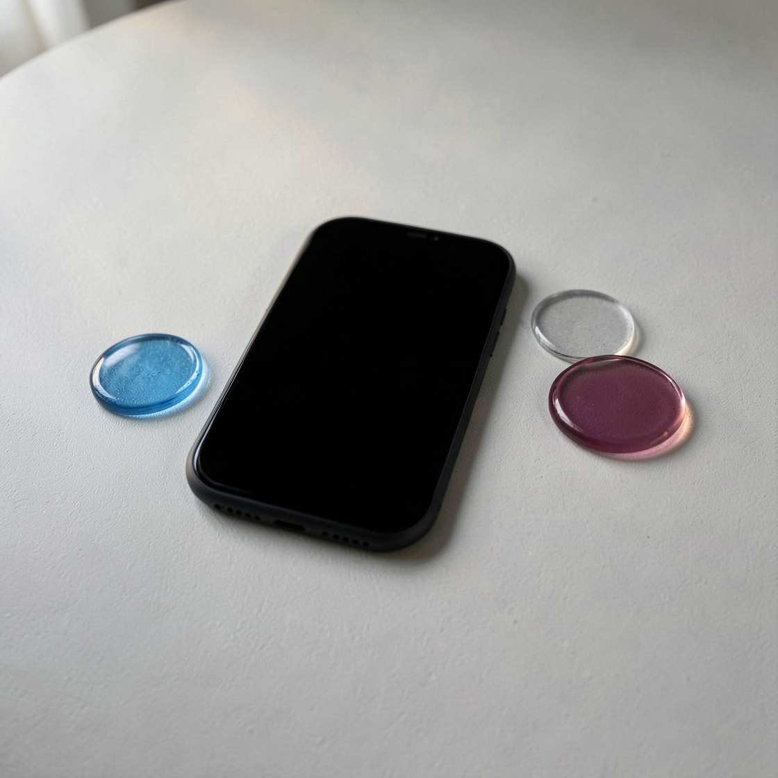 Black-screen smartphone beside blue, magenta and clear glass discs on a pale desk