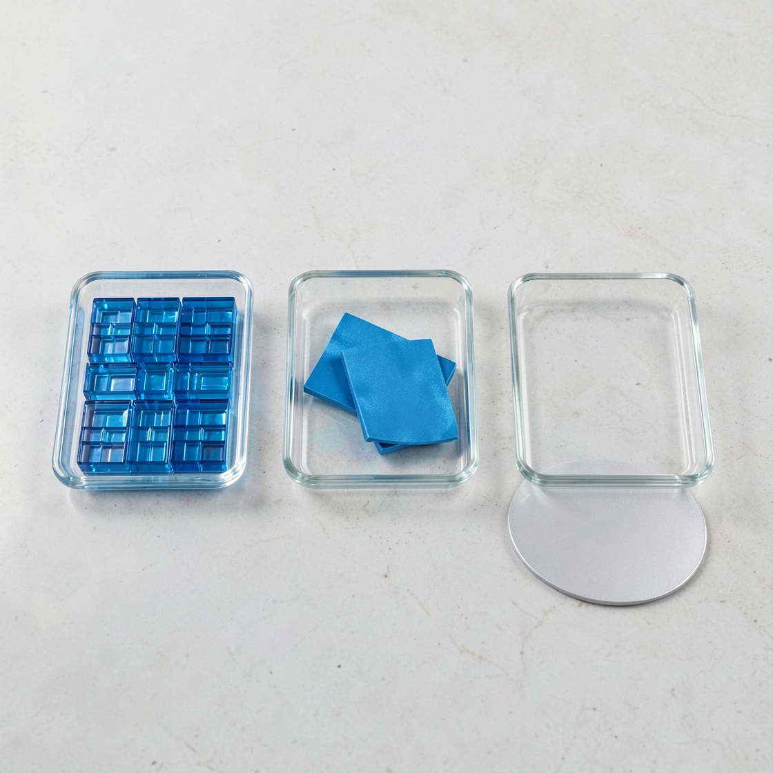 Three glass trays with blue pieces and an unmarked silver disc on a pale tabletop