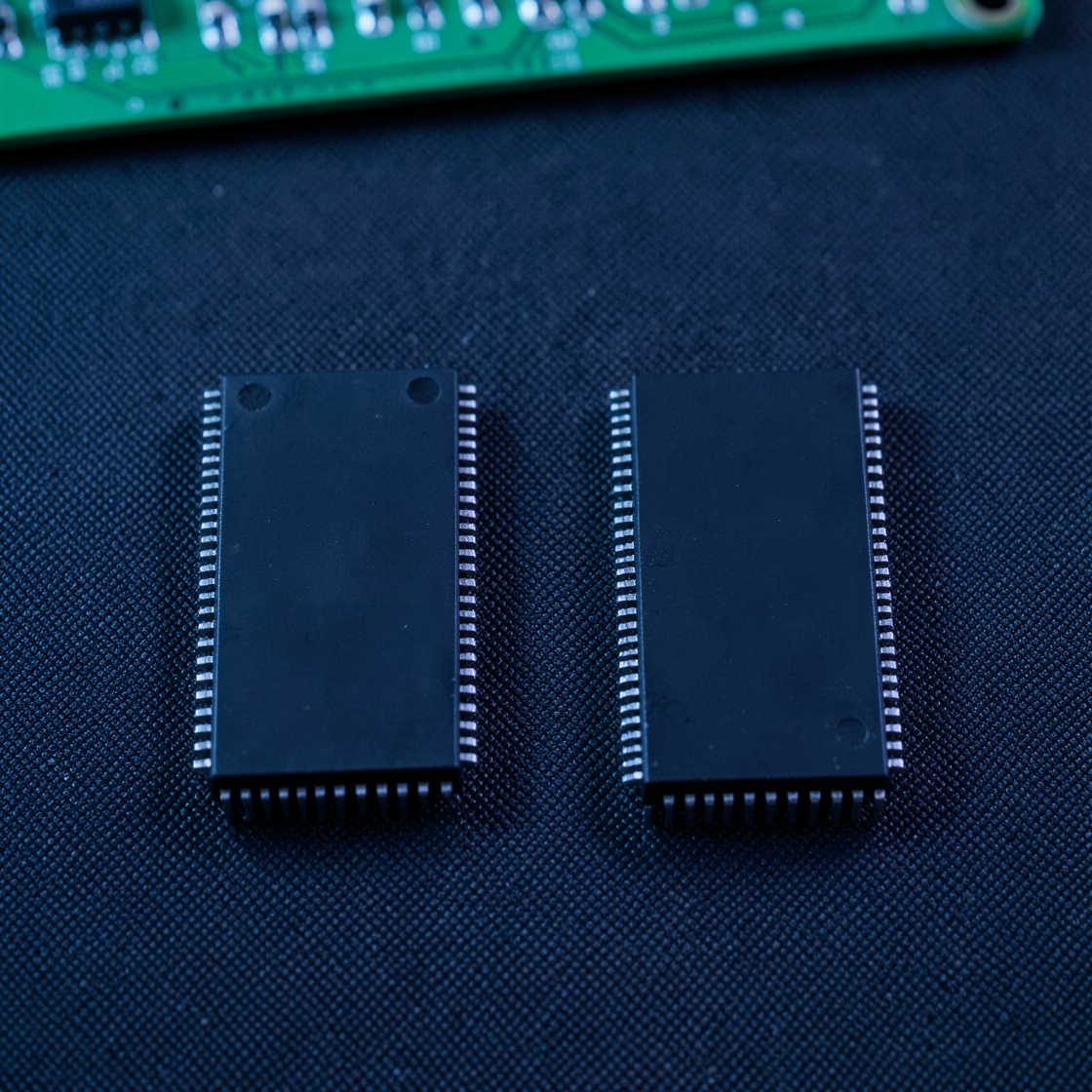 Two unmarked rectangular integrated circuits on a dark mat beside a blurred green circuit board.