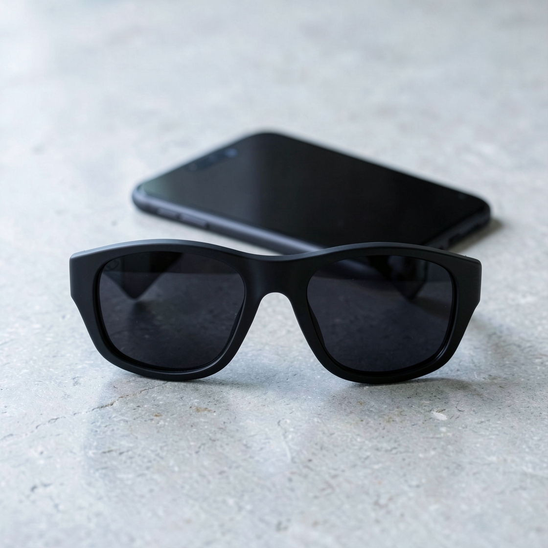 Plain black sunglasses with dark lenses on a light stone surface, with a dark-screen phone behind them.