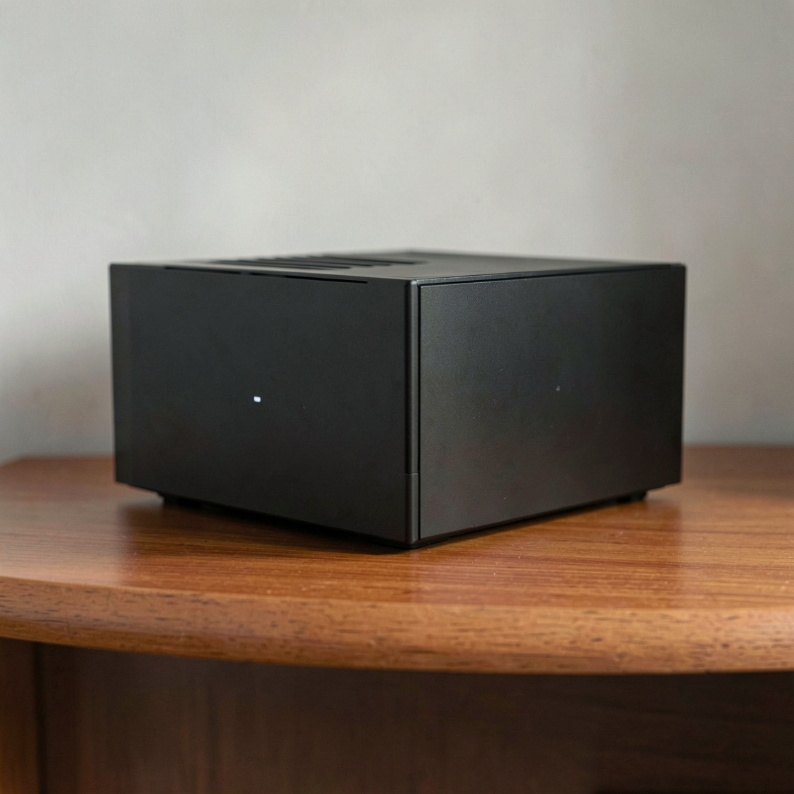A plain compact black computer enclosure with cooling slots and a small white light on a wooden shelf.