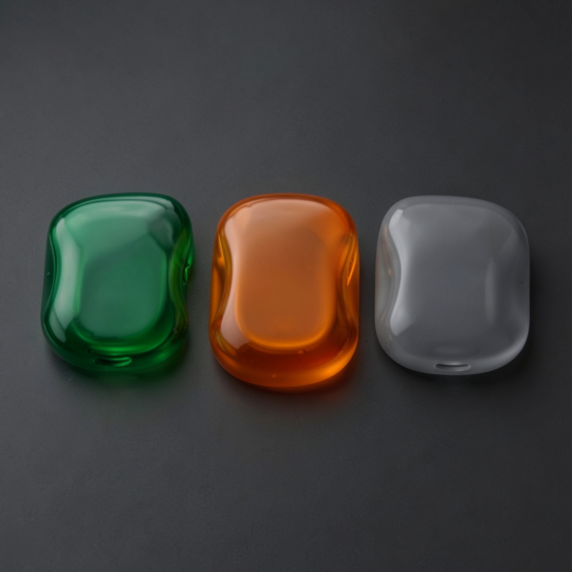 Three smooth rounded translucent plastic forms in green, orange and smoke gray on a dark surface.