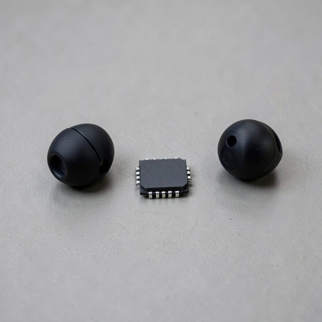 Two unbranded black wireless earbuds flanking a blank microchip on a light gray surface